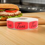 Tuna sticker red identification label with black print on a roll of stickers with a tuna fish sandwich in background